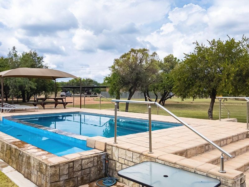 An elevated swimming pool area featuring stone cladding, stainless steel safety railings, and a paved deck with a shade sail in a rural setting.