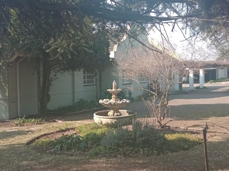 A view of the rear garden featuring a central stone fountain, mature trees, and a covered walkway leading to the main building structure.