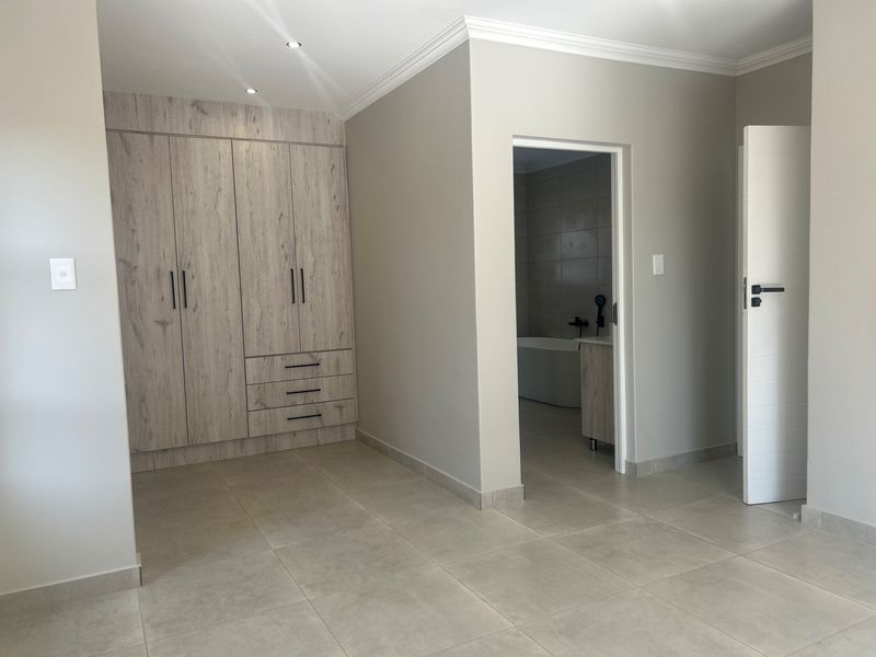 A clean, modern bedroom featuring neutral tiled floors, ample built-in cupboards with wood-grain finish, and direct access to an en-suite bathroom.