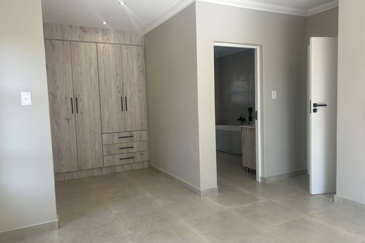 A clean, modern bedroom featuring neutral tiled floors, ample built-in cupboards with wood-grain finish, and direct access to an en-suite bathroom.