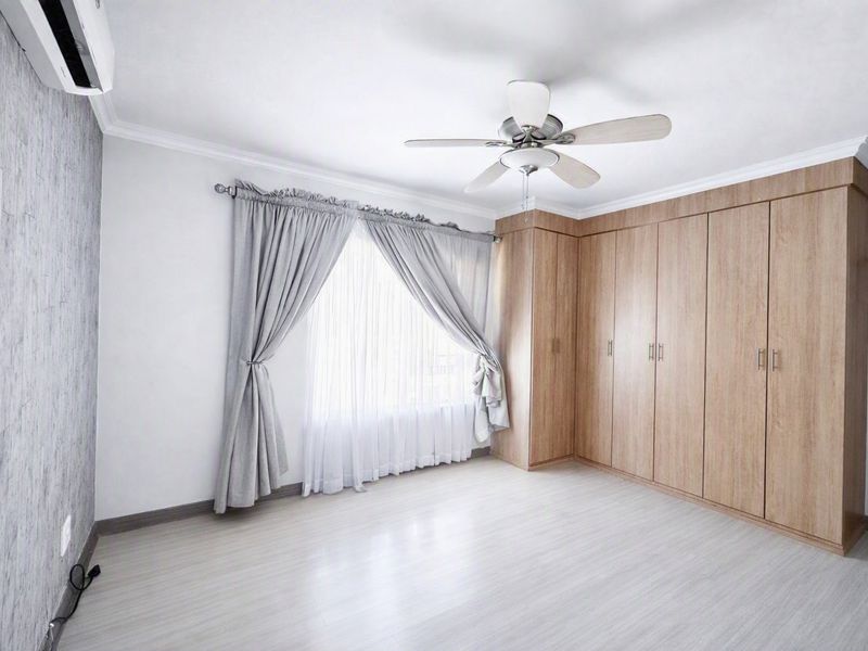 A bright, empty bedroom featuring light-toned laminate flooring, extensive built-in wooden cupboards, a ceiling fan, and a wall-mounted air conditioning unit.