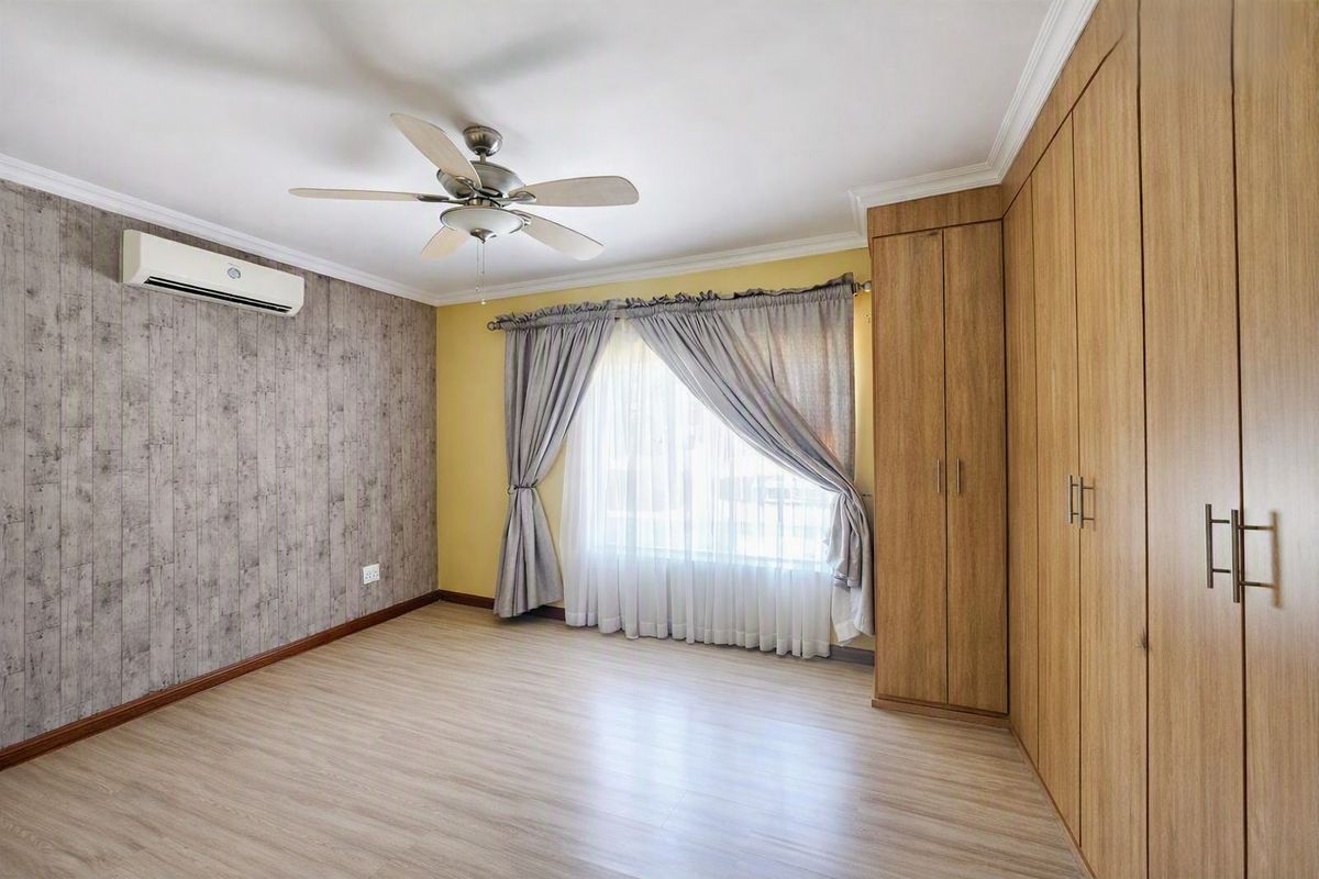 A bright, empty bedroom featuring laminate flooring, built-in wooden cupboards, a ceiling fan, and a wall-mounted air conditioning unit.