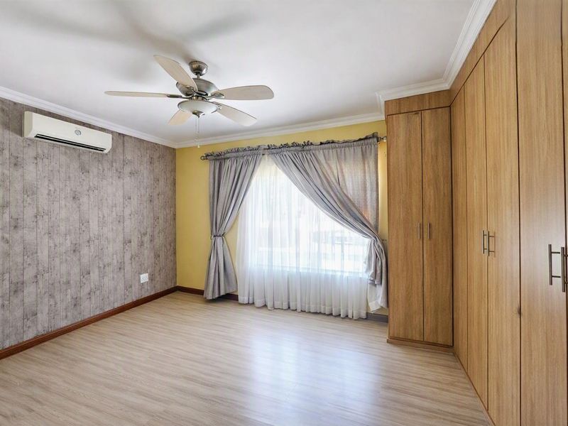 A bright, empty bedroom featuring laminate flooring, built-in wooden cupboards, a ceiling fan, and a wall-mounted air conditioning unit.