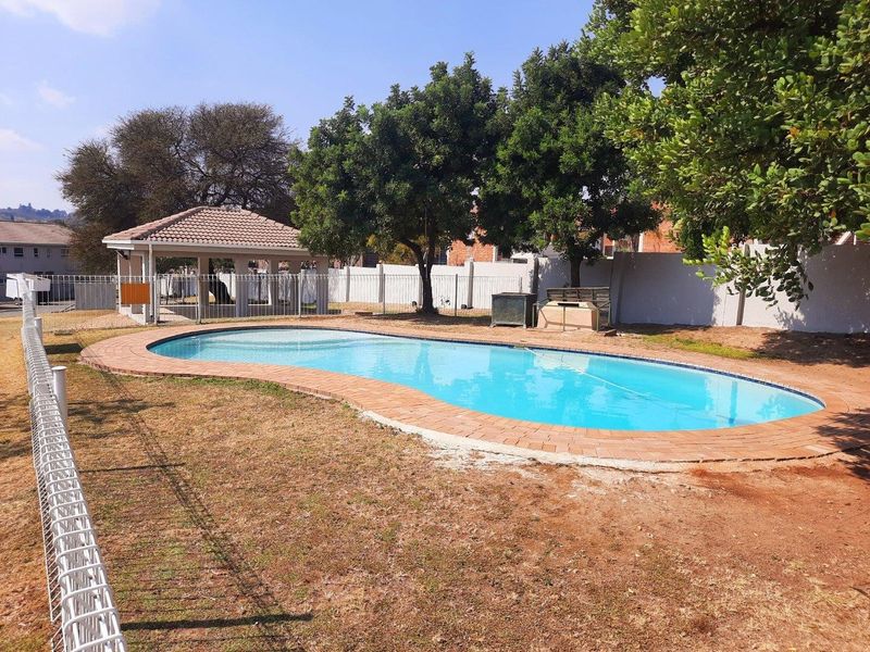 Communal swimming pool area featuring a paved surround, a covered gazebo, and perimeter fencing within a landscaped complex setting.