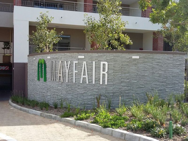 The entrance to the Mayfair apartment complex featuring a modern stone-clad signage wall, landscaped garden, and secure driveway access.