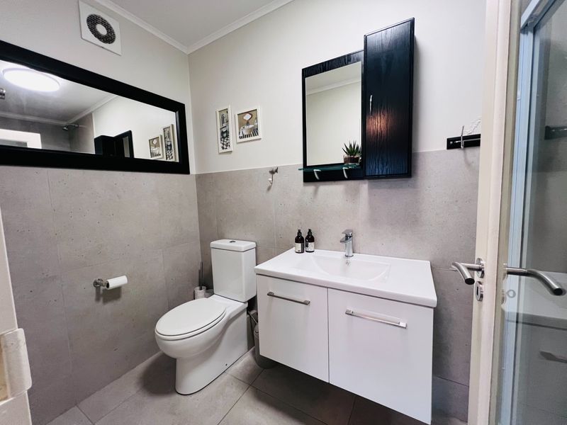 A modern bathroom featuring a white vanity cabinet, ceramic basin, wall-mounted medicine cabinet, large mirror, and neutral wall and floor tiling.