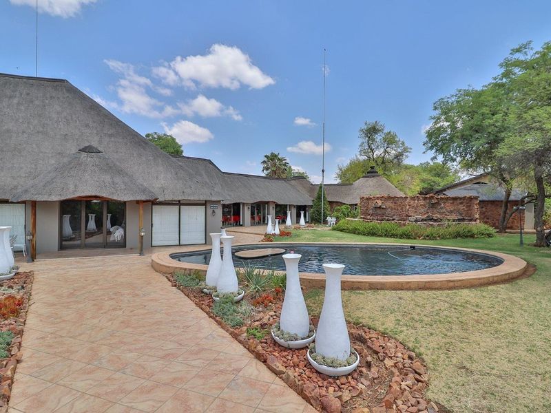 A well-maintained outdoor pool area featuring a circular swimming pool, stone retaining wall, and a paved walkway set against a thatched-roof farm building.