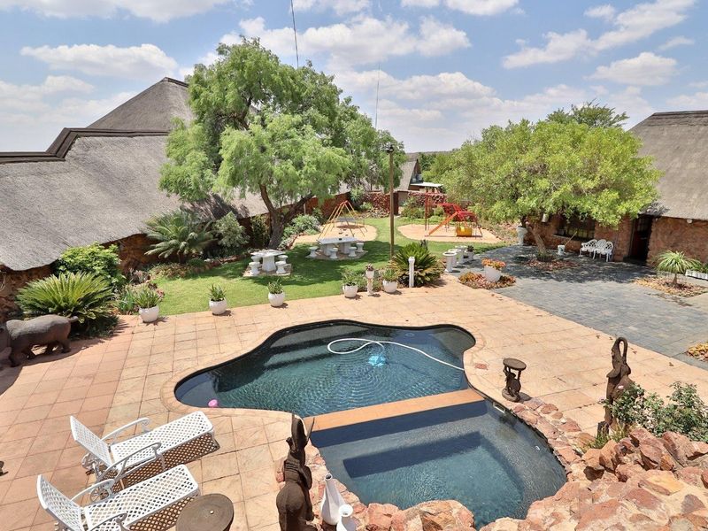A spacious outdoor entertainment area featuring a swimming pool, paved patio, landscaped garden, and traditional thatched roof structures.