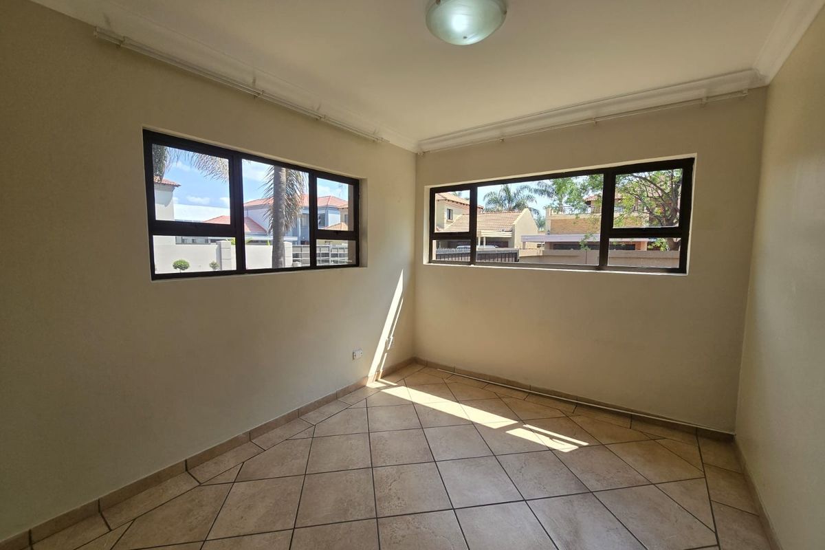 A bright, empty bedroom featuring neutral walls, tiled flooring, and large aluminium-framed windows providing natural light.