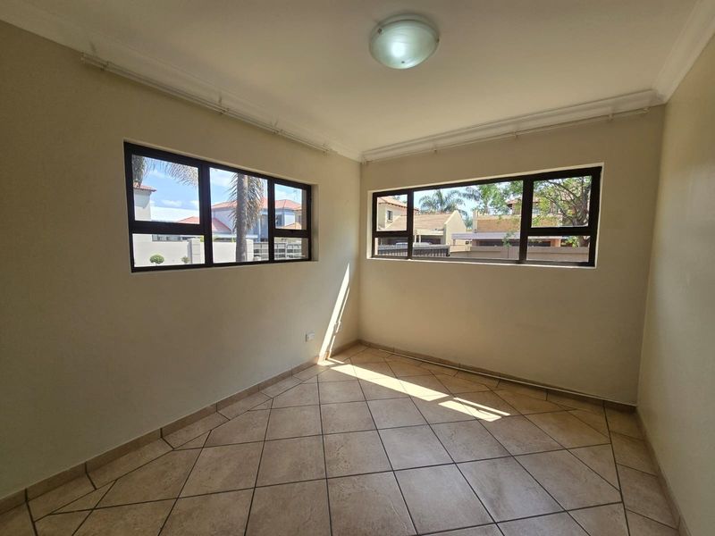 A bright, empty bedroom featuring neutral walls, tiled flooring, and large aluminium-framed windows providing natural light.