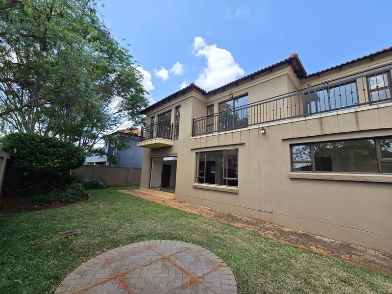 Rear view of a two-story house featuring a paved patio, upper-level balcony with wrought iron railings, and large windows overlooking a private garden.