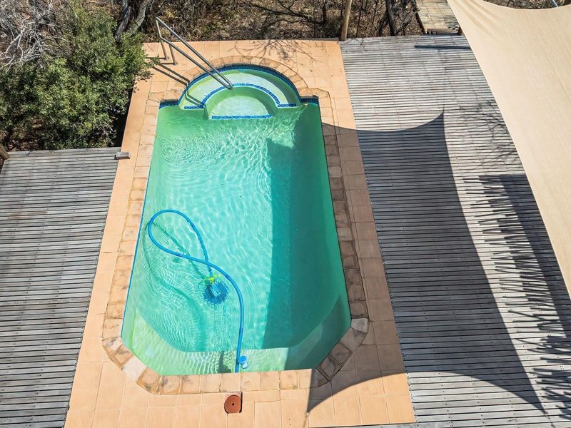 An elevated view of a swimming pool surrounded by timber decking, featuring a shade sail and integrated pool steps with a metal handrail.