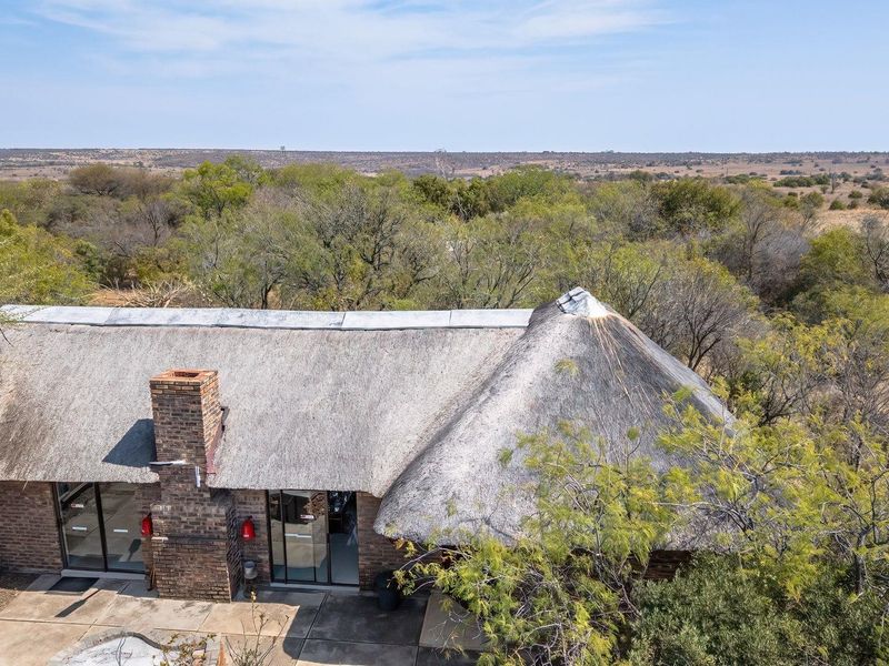 Aerial view of a farm property featuring a traditional thatch roof, brick chimney, and a paved patio area surrounded by natural landscape.