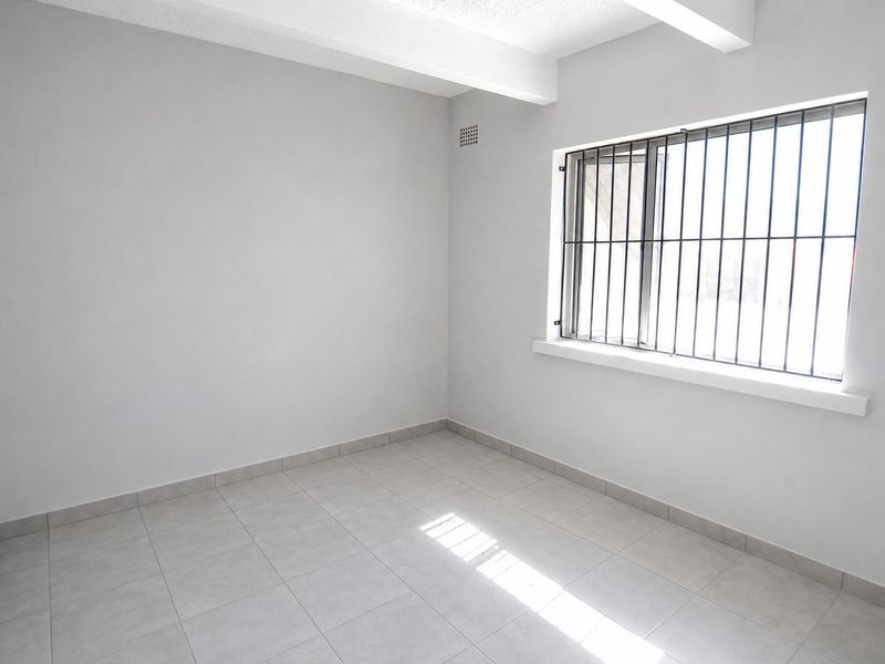 A clean, empty bedroom featuring neutral tiled flooring, a window with security bars, and a simple, bright interior finish.