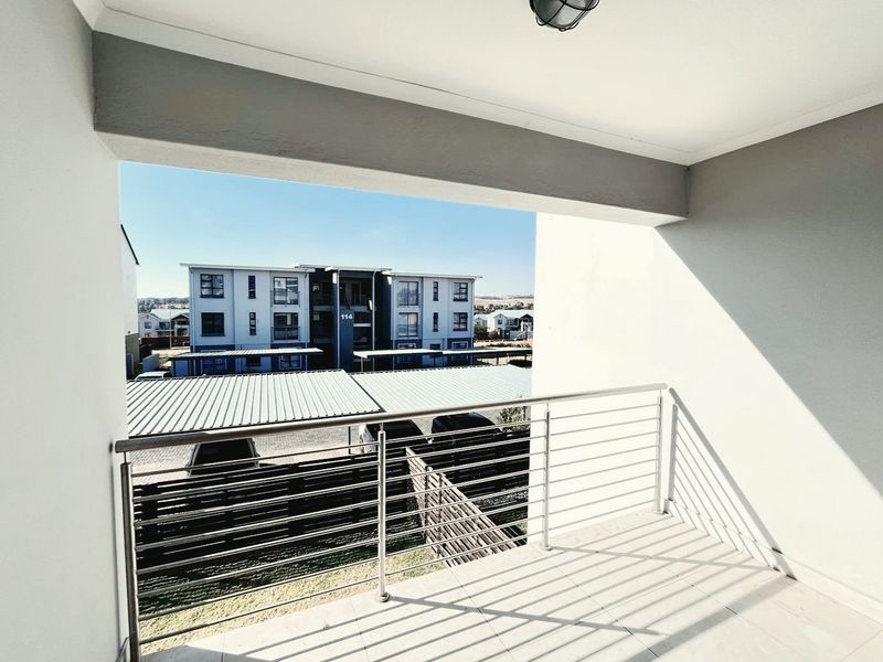 A clean, covered balcony featuring tiled flooring and a modern stainless steel balustrade, offering an elevated view of the surrounding residential complex.