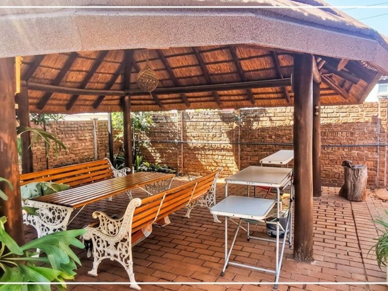 A covered outdoor patio area featuring a traditional thatch roof, timber support posts, and brick paving, enclosed by a face brick boundary wall.