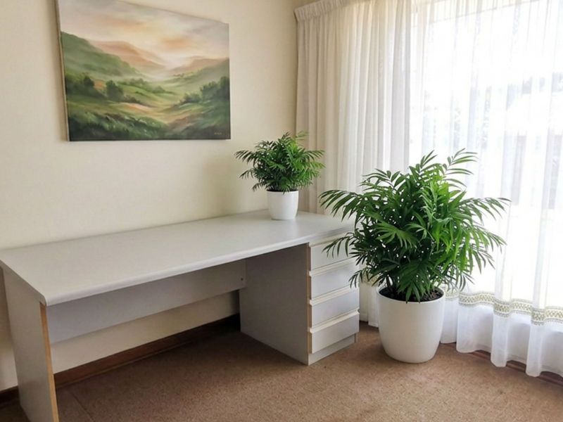 A functional home office space featuring a built-in desk, neutral carpeting, and large windows with sheer curtains allowing for natural light.