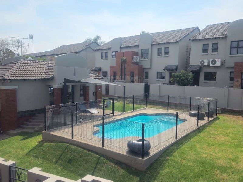 A well-maintained communal pool area featuring a fenced swimming pool, paved surround, and landscaped lawn within a residential apartment complex.