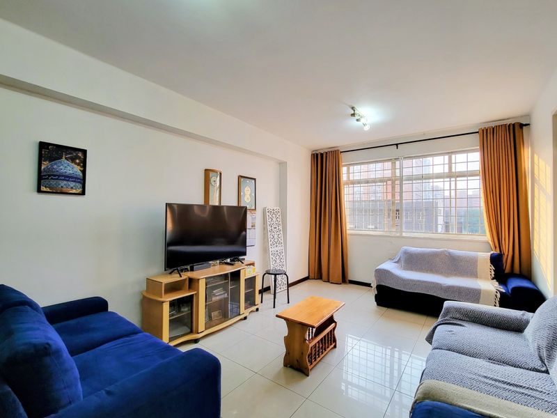 A bright living room featuring large windows with security bars and neutral tiled flooring, offering a functional and well-lit space.