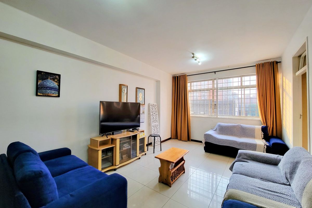 A bright living room featuring large windows with security bars and neutral tiled flooring, offering a functional and well-lit space.