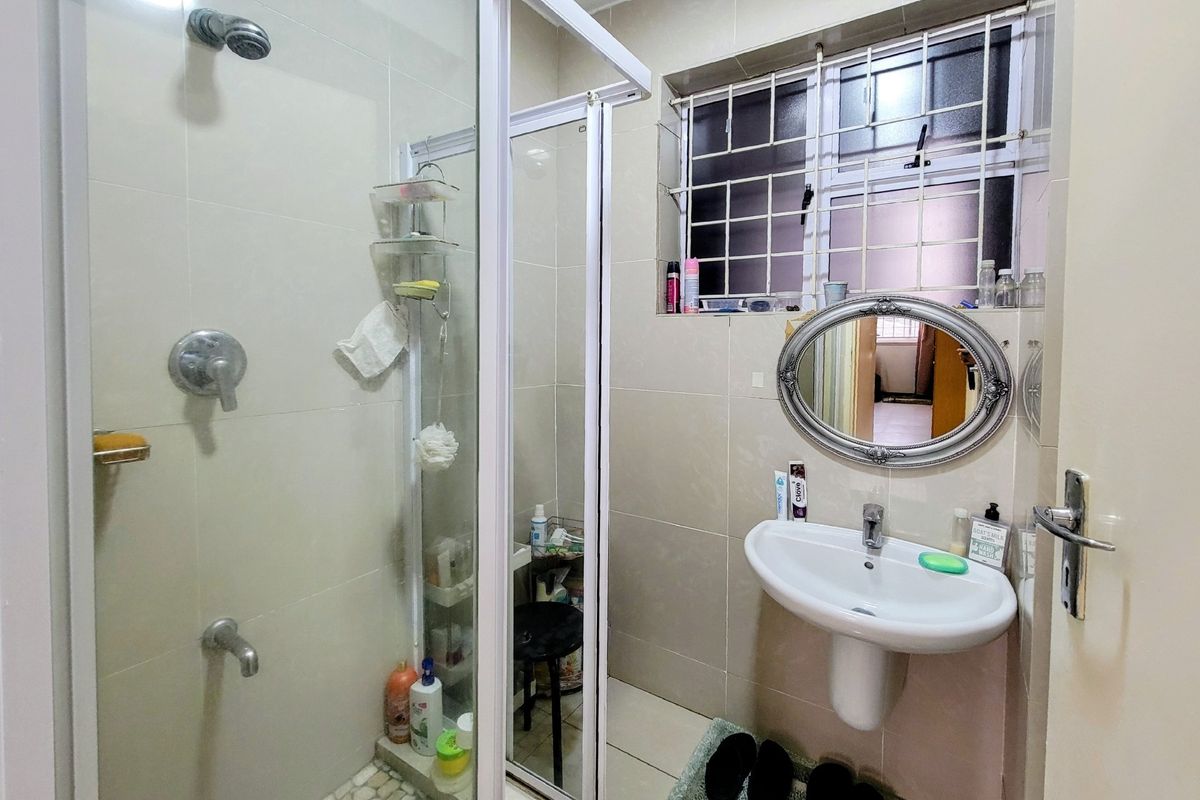 A functional bathroom featuring a glass-enclosed shower, pedestal basin, and neutral wall tiling with security bars on the window.