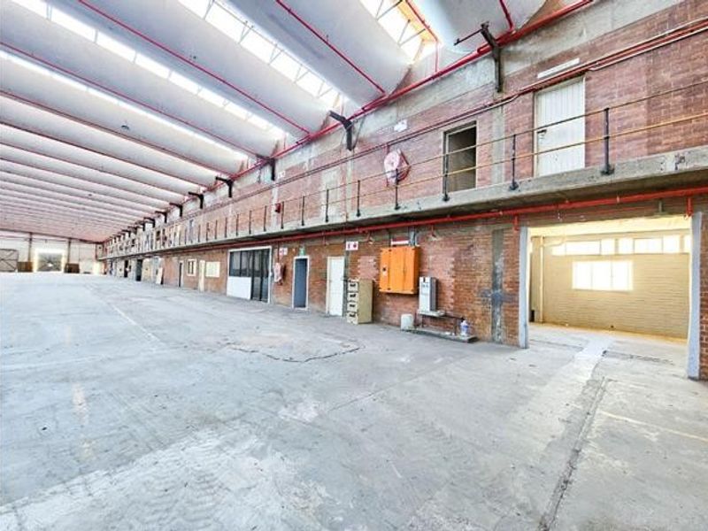 Expansive industrial warehouse space featuring high ceilings with skylights, exposed brick walls, concrete flooring, and a mezzanine level with safety railings.