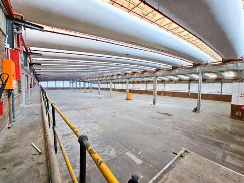 Expansive industrial warehouse space featuring high ceilings with exposed ventilation ducting, concrete flooring, and robust steel structural elements.