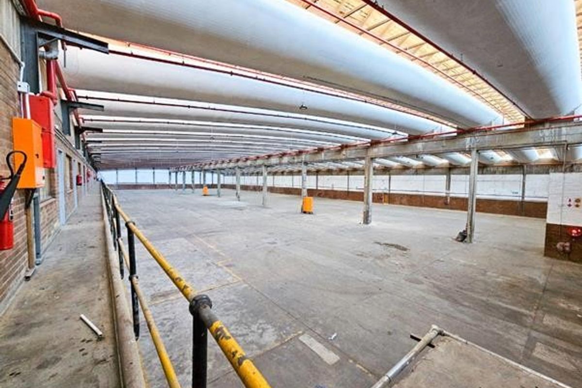 Expansive industrial warehouse space featuring high ceilings with exposed ventilation ducting, concrete flooring, and robust steel structural elements.