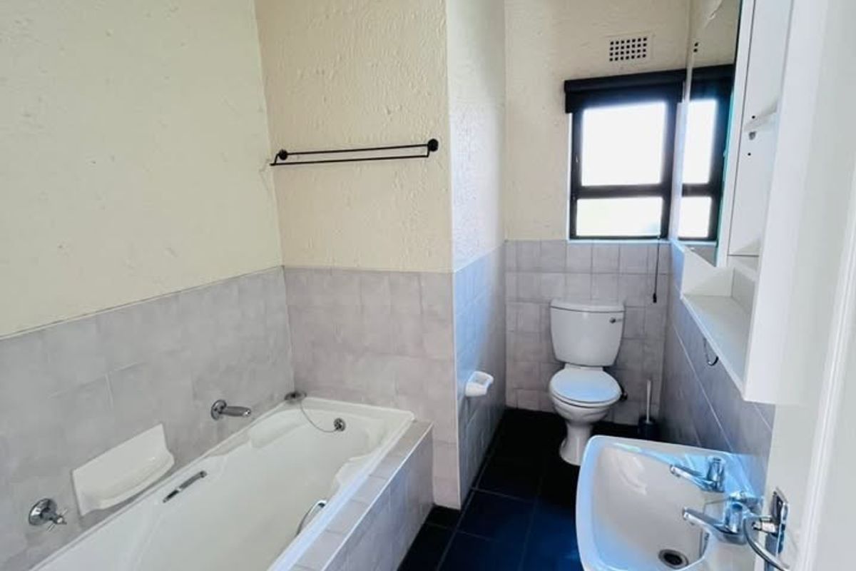 A functional bathroom featuring a bathtub, pedestal sink, toilet, and dark tiled flooring, complemented by exposed ceiling beams and neutral wall finishes.