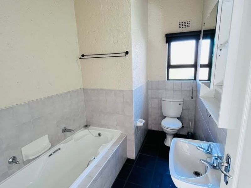 A functional bathroom featuring a bathtub, pedestal sink, toilet, and dark tiled flooring, complemented by exposed ceiling beams and neutral wall finishes.