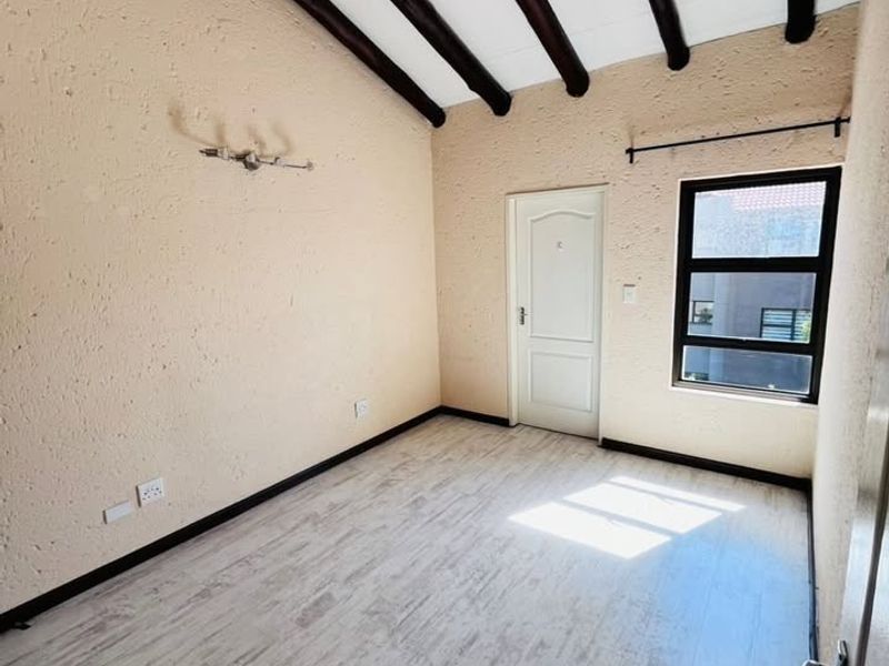 A bright, empty bedroom featuring exposed timber ceiling beams, textured walls, and light-toned laminate flooring with a window providing natural light.