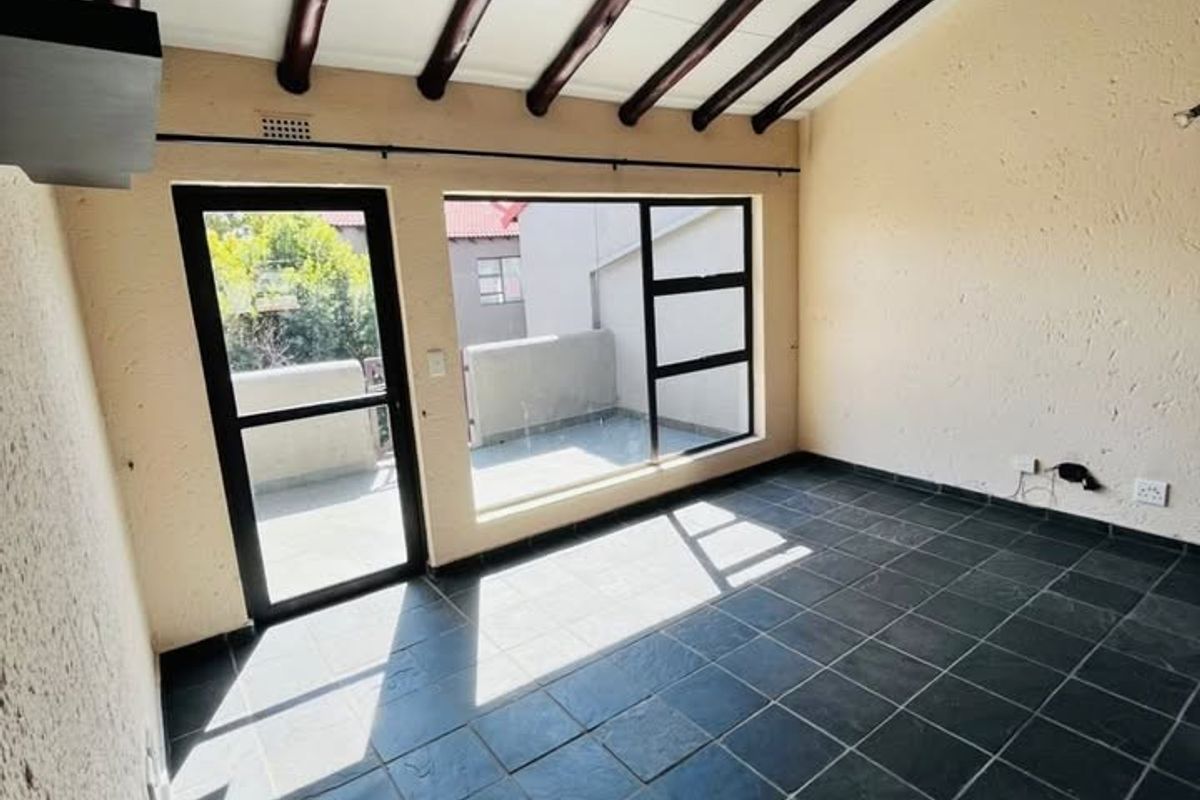 An empty living area featuring exposed timber ceiling beams, dark slate tile flooring, and direct access to an outdoor balcony through a glass door.