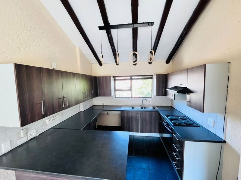A well-lit kitchen featuring dark wood cabinetry, contrasting countertops, an electric hob with extractor, and distinctive exposed timber ceiling beams.