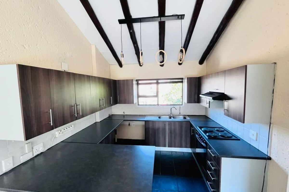 A well-lit kitchen featuring dark wood cabinetry, contrasting countertops, an electric hob with extractor, and distinctive exposed timber ceiling beams.