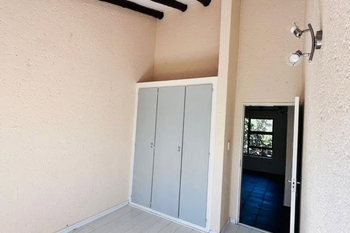 A bright, empty bedroom featuring exposed timber ceiling beams, built-in wardrobe storage, textured walls, and light-toned tiled flooring.