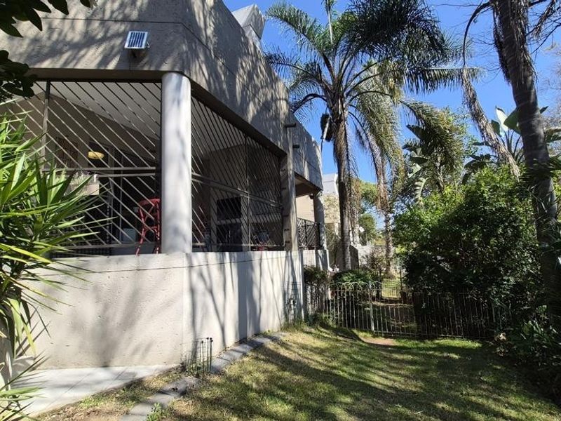 Rear view of the apartment featuring a covered patio area with security bars, overlooking a private garden space with mature trees.