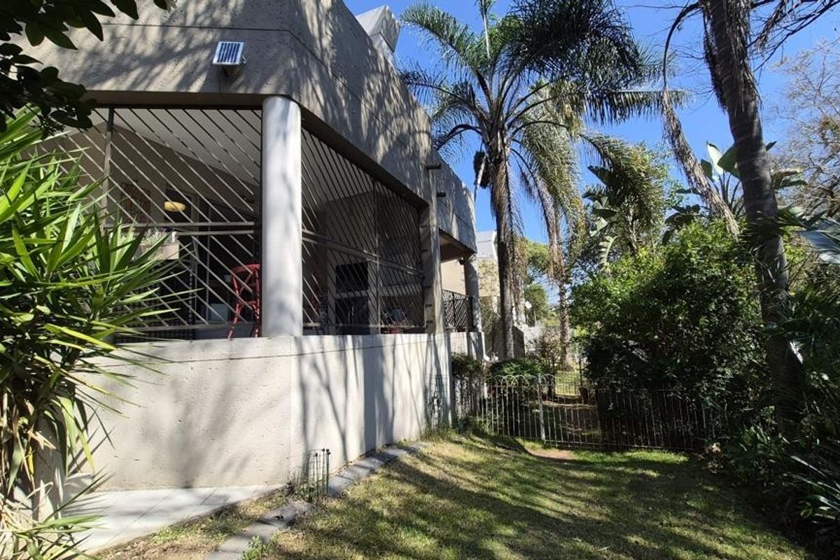 Rear view of the apartment featuring a covered patio area with security bars, overlooking a private garden space with mature trees.