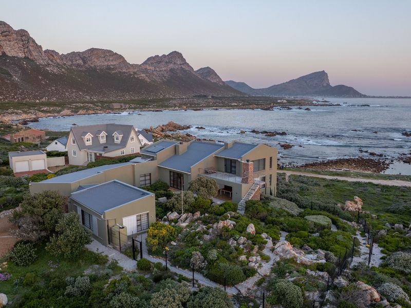 Aerial view of a coastal property featuring a multi-level structure with metal roofing, brick chimney, and balcony, set against a scenic ocean and mountain backdrop.