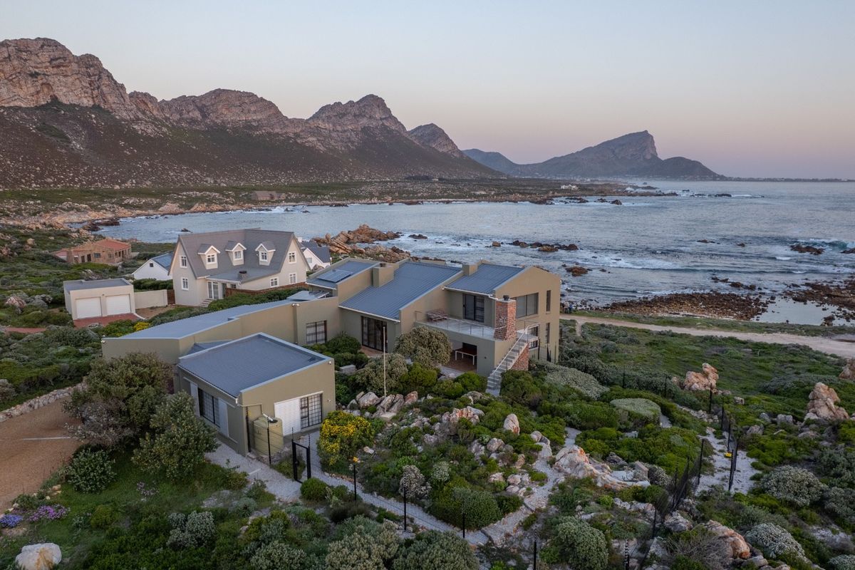 Aerial view of a coastal property featuring a multi-level structure with metal roofing, brick chimney, and balcony, set against a scenic ocean and mountain backdrop.