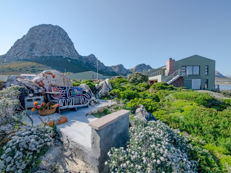 A scenic rear garden area featuring natural rock landscaping, a concrete seating platform, and views of the modern residence and surrounding mountains.