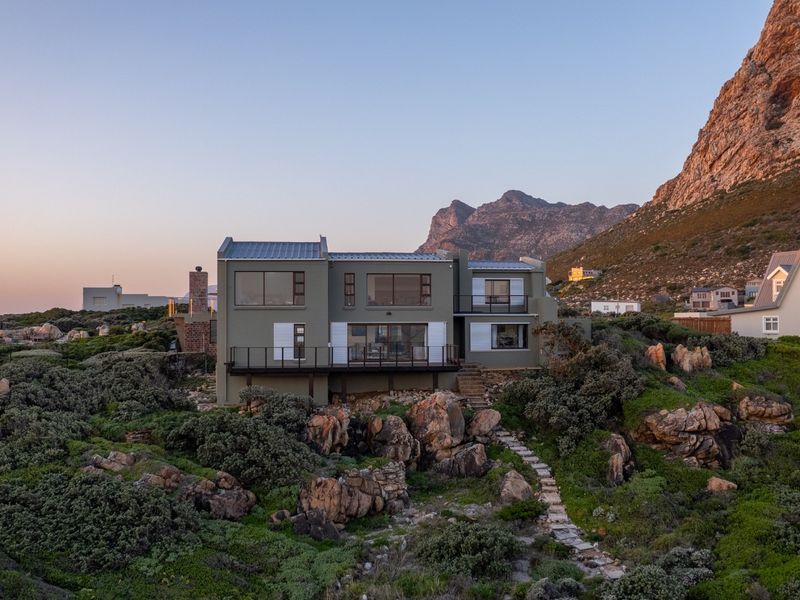 A stunning multi-level coastal home featuring a metal roof, expansive windows, and a balcony, set against a dramatic mountain backdrop with natural rocky landscaping.