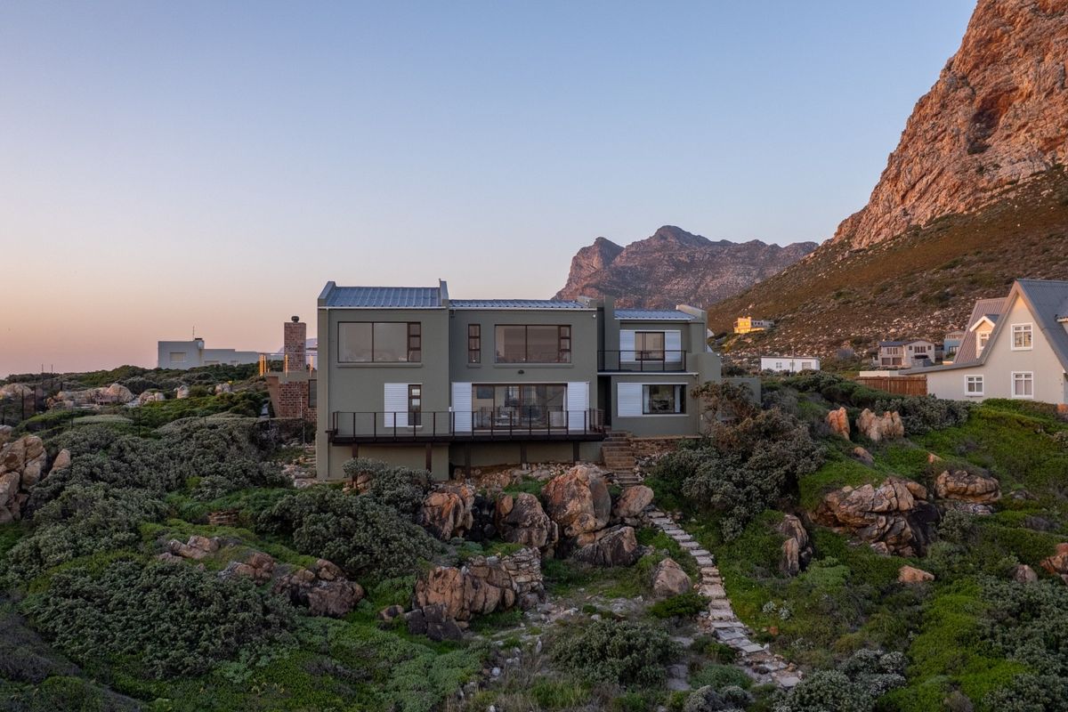 A stunning multi-level coastal home featuring a metal roof, expansive windows, and a balcony, set against a dramatic mountain backdrop with natural rocky landscaping.