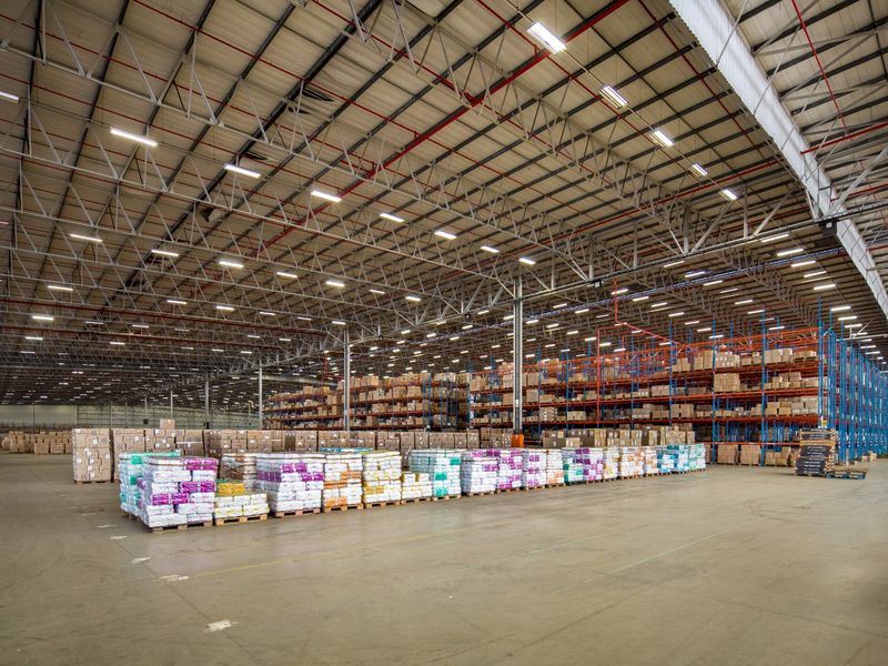 A large industrial warehouse space featuring high ceilings with exposed steel trusses, industrial lighting, sprinkler systems, and extensive pallet racking for storage.