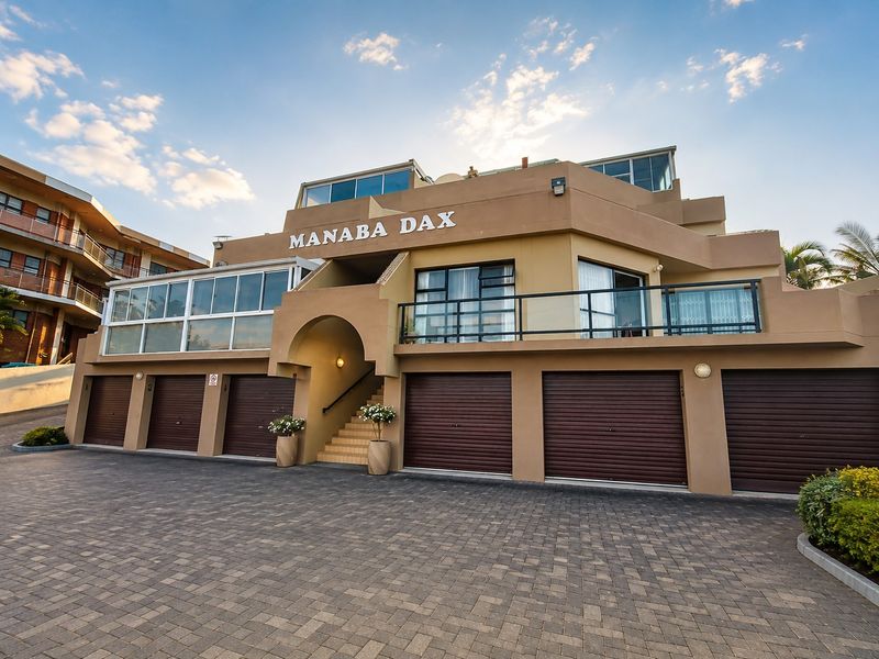 Front elevation of the Manaba Dax apartment complex featuring multiple garage units, a paved driveway, and upper-level balconies with glass balustrades.
