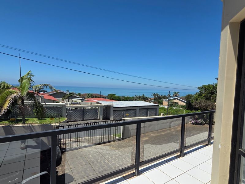 A balcony featuring a glass balustrade and tiled flooring, offering an elevated view of the surrounding neighborhood and the ocean in the distance.