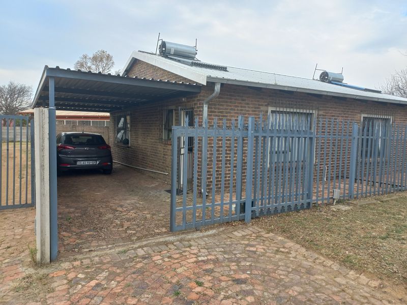 Front exterior view of a brick apartment featuring a carport, paved driveway, security fencing, and solar water heating system.