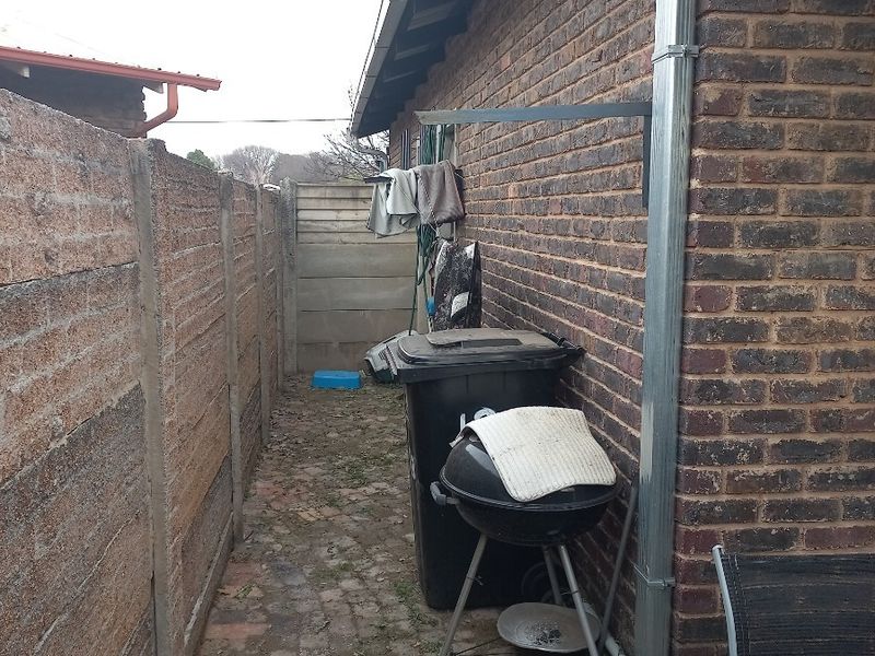 A narrow rear exterior side passage featuring brick walling, a metal downpipe, and paved ground surface.