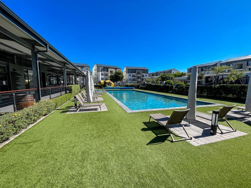 A well-maintained communal swimming pool area featuring artificial turf, paved decking, and a covered walkway, set within a modern apartment complex.