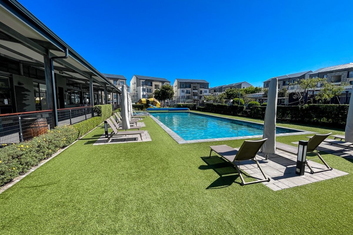 A well-maintained communal swimming pool area featuring artificial turf, paved decking, and a covered walkway, set within a modern apartment complex.