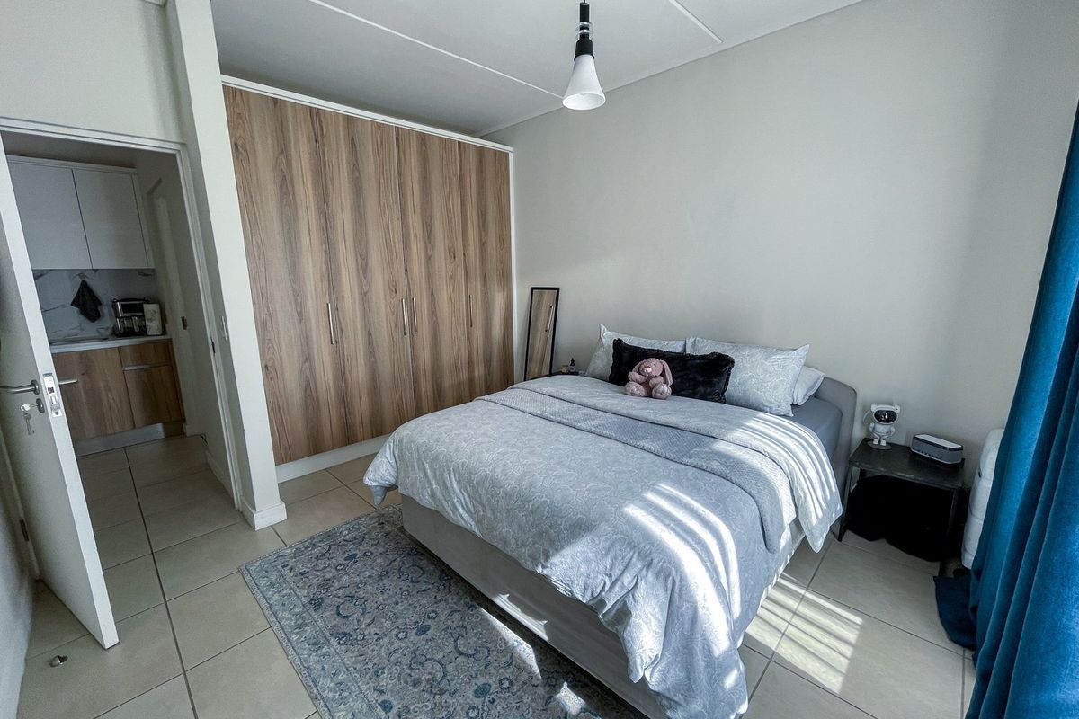 A well-maintained bedroom featuring built-in wood-finish cupboards, neutral tiled flooring, and a pendant light fixture, with an adjacent view into the kitchen area.